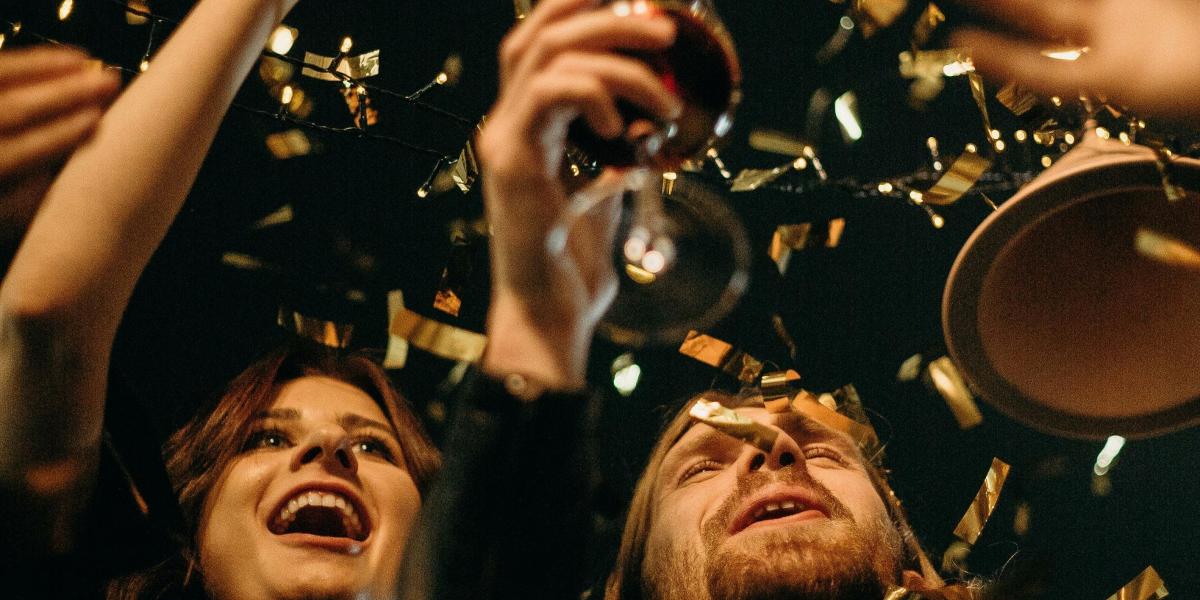 People celebrating with wine glasses raised, smiling amid falling gold confetti at a holiday party.