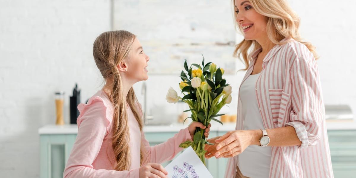 Cheerful kid offering flowers and a card to her mother. 
